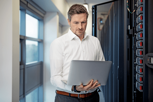 Ein IT-Techniker steht mit seinem Laptop im Serverraum seines Kunden und prüft die Funktionsfähigkeit der Geräte.